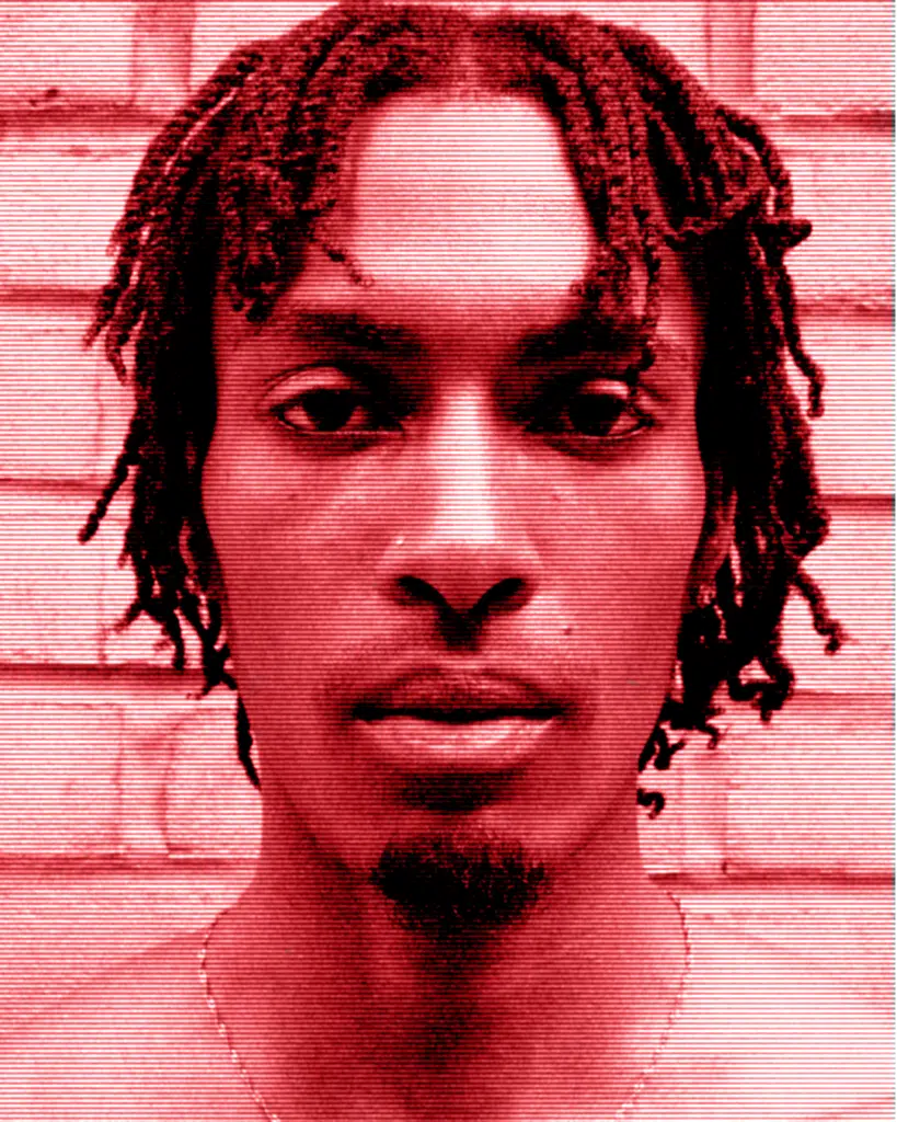 A man with dreadlocks is standing in front of a brick wall.