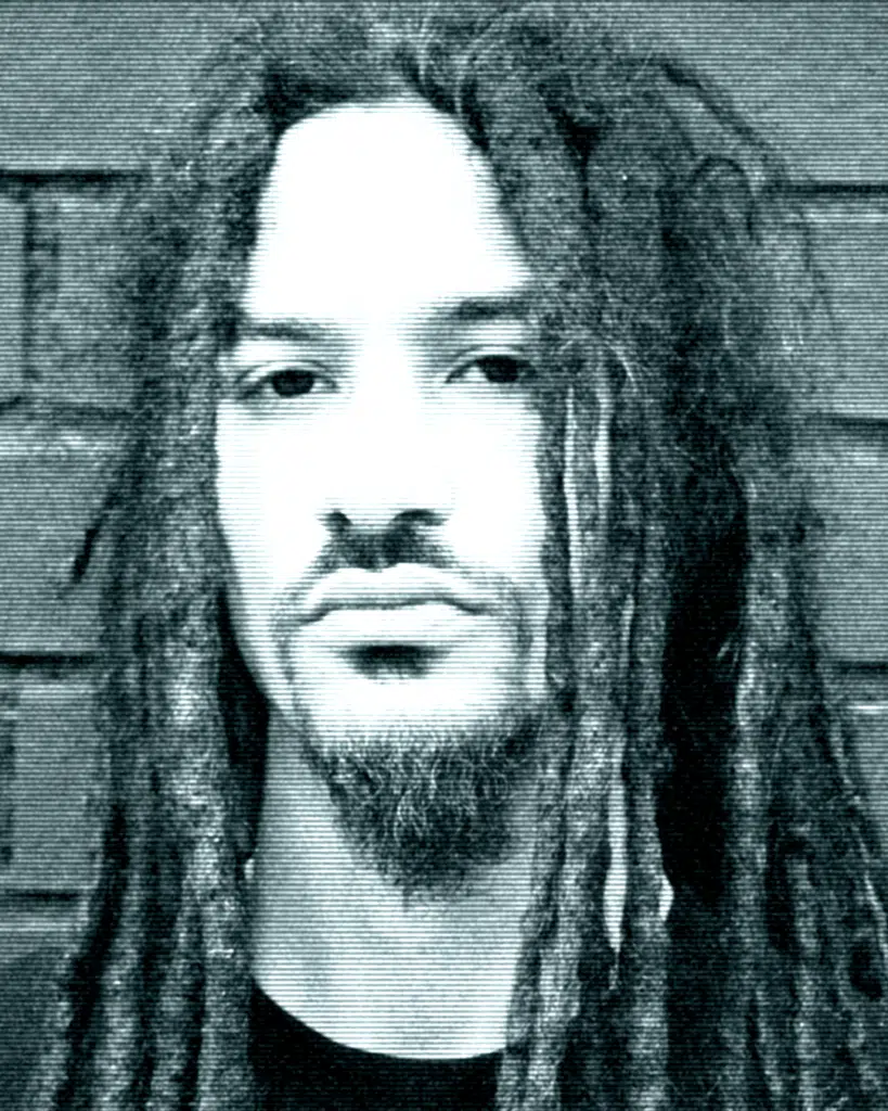 A man with dreadlocks and beard standing in front of a brick wall.