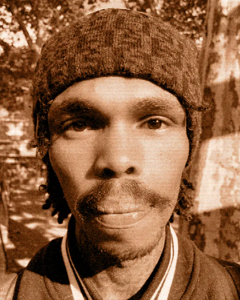 A man with a beard and hat in front of trees.