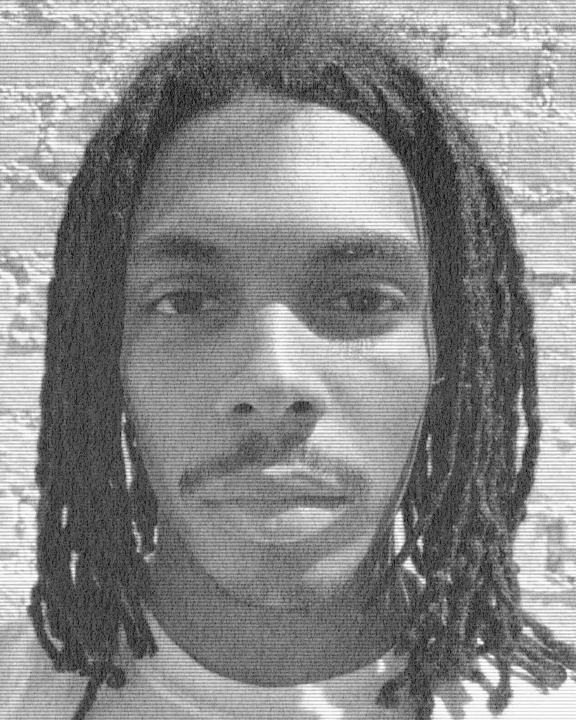 A man with dreadlocks is standing in front of a brick wall.