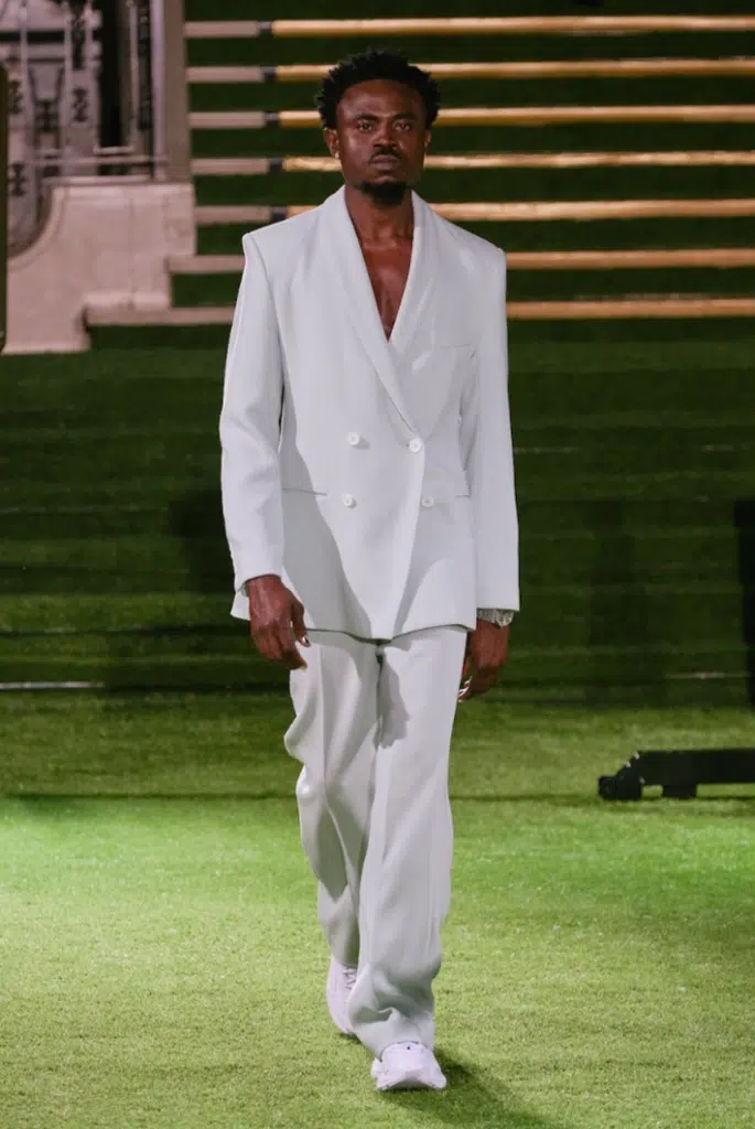 A man in white suit and tie standing on grass.