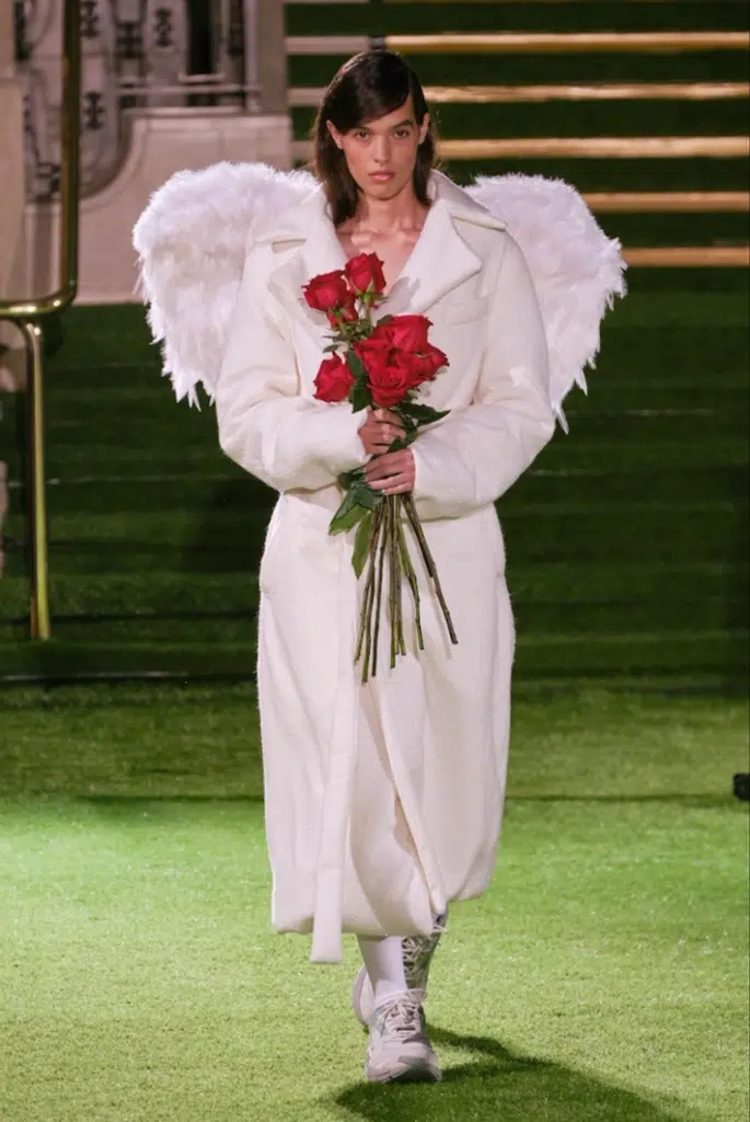 A woman in white robe holding roses and wearing angel wings.