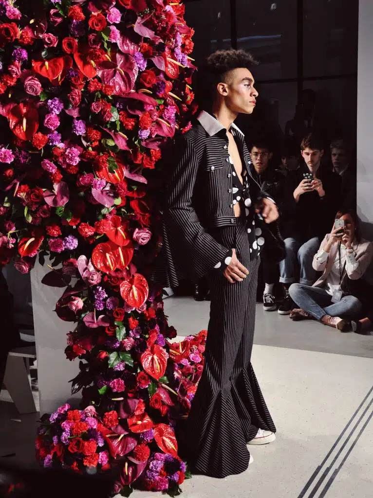 A man in black suit and tie standing next to flowers.