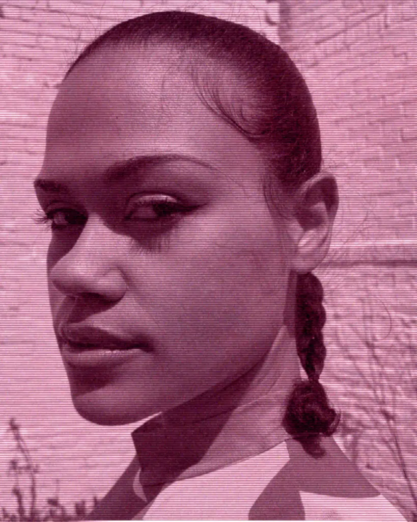 A woman with braids is standing in front of a brick wall.